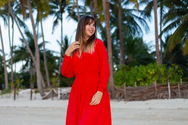 Кавказская женщина в красном летнем платье в романтическом счастливом настроении на тропическом пляже с белым песком на закате