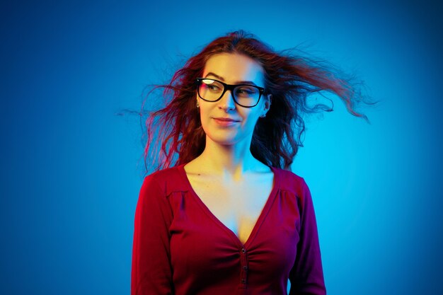 Ritratto di donna caucasica isolato su sfondo blu studio in luce al neon. bellissimo modello femminile con i capelli rossi in casual. concetto di emozioni umane, espressione facciale, vendite, annuncio. guardando di lato.