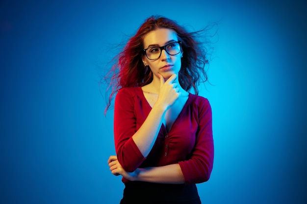 Caucasian woman's portrait isolated on blue studio background in neon light. Beautiful female model with red hair in casual style. Concept of human emotions, facial expression, sales, ad. Thoughtful.