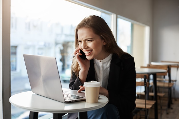 白人女性がカフェに座っている、コーヒーを飲む、スマートフォンで話している、笑顔でノートパソコンの画面を見て