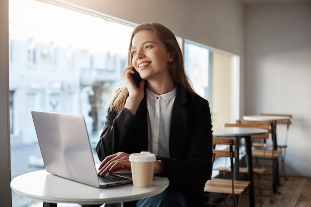 caucasian woman sitting in cafe with laptop, drinking coffee, talking on smartphone