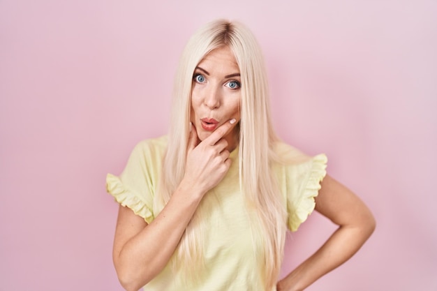 Free photo caucasian woman standing over pink background looking fascinated with disbelief surprise and amazed expression with hands on chin