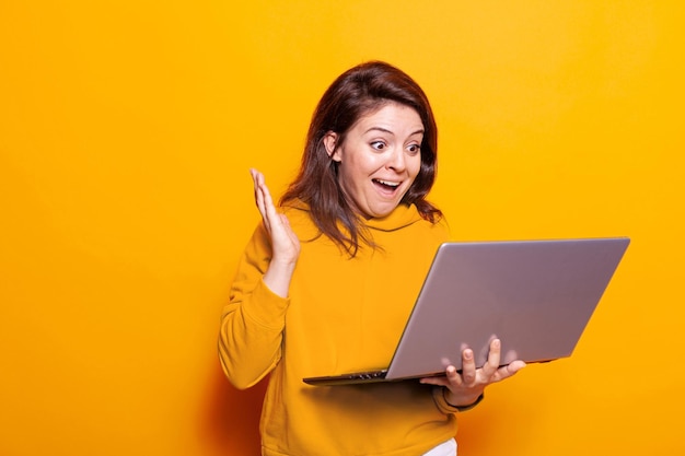 Donna caucasica agitando la mano alla videochiamata sul laptop in studio. giovane che si sente eccitato e utilizza la videoconferenza online mentre tiene in mano un dispositivo moderno con webcam. conversazione a distanza