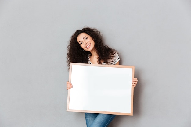 Caucasian woman with beautiful hair posing over grey wall holding piece of art in hands expressing admiration about portrait copy space