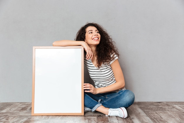 Free photo caucasian woman with beautiful hair posing with legs crossed demonstrating big great painting or portrait isolated over grey wall copy space