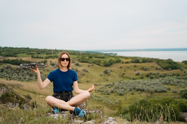 A Caucasian woman with a drone in her hand, sitting on a green rocky hill with sky