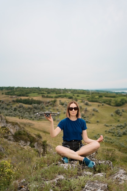 A Caucasian woman with a drone in her hand, sitting on a green rocky hill with sky