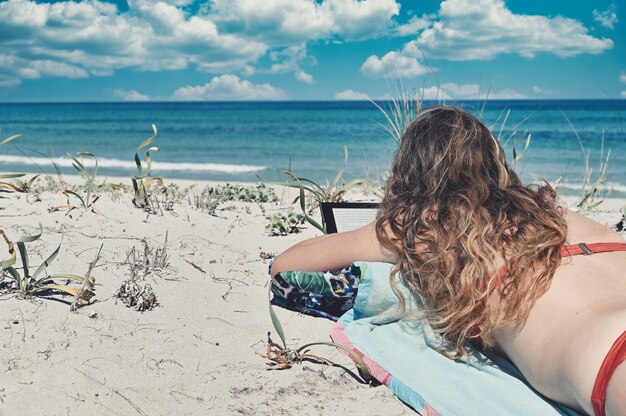 Caucasian woman with long hair, wearing a red bikini, lying on the beach next to the blue sea