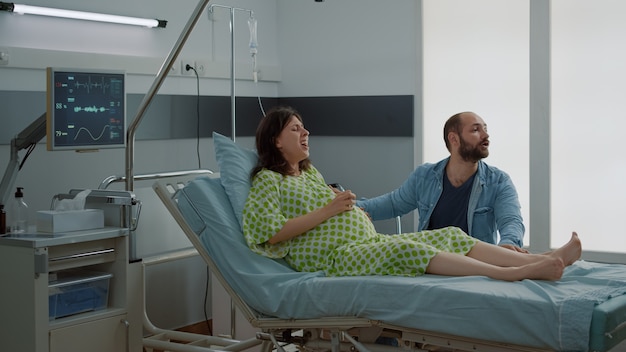 Foto gratuita donna caucasica con gravidanza con contrazioni dolorose con il marito che tiene la mano. infermiera afroamericana che sostiene il parto in corsia ospedaliera. coppia in attesa di un bambino