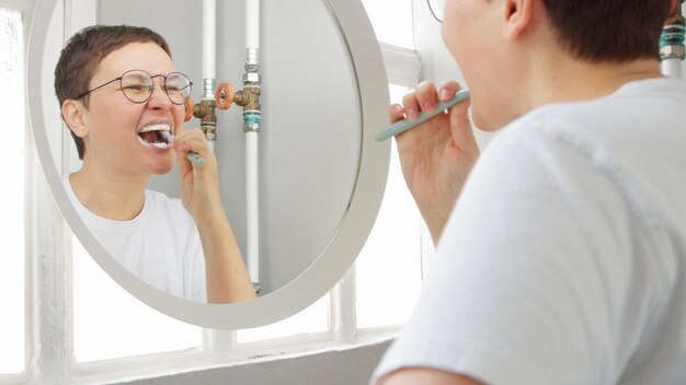 日光に満ちたバスルームの鏡の前で短いヘアカットをした白人女性が歯を磨く