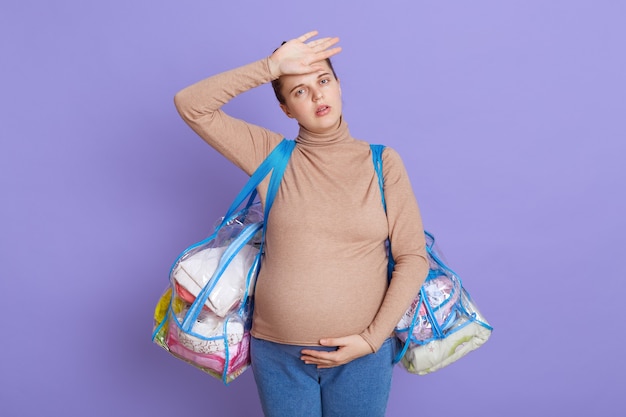 Caucasian young beautiful pregnant, tired future mom feeling fatigue and headache, looks tired and exhausted, touching her forehead, holding two bags, going to maternity house.