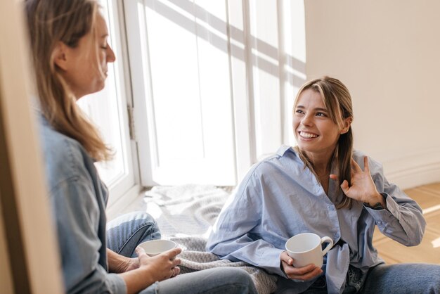 Caucasian young girlfriends enjoy weekend together, drink coffee and talk at window home. Beauty, people emotions and vacation concept.