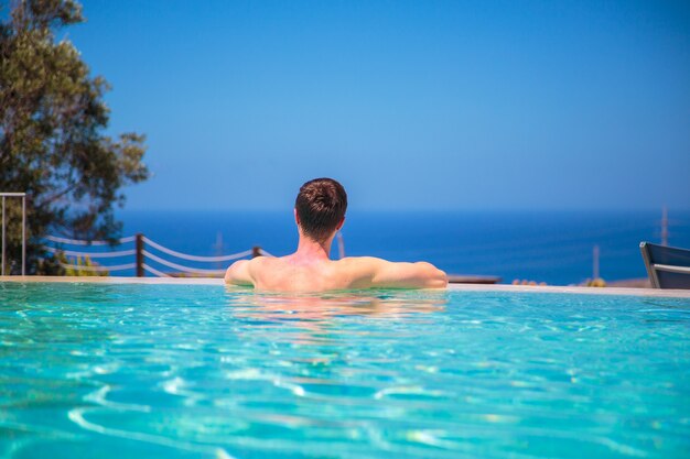 Кавказский молодой человек в пейзажном бассейне, наблюдая за видом на океан, расслабляясь и наслаждаясь своей жизнью