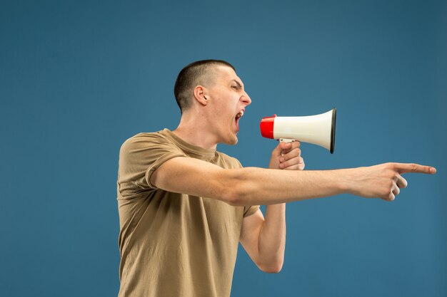 Caucasian young man's half-length portrait on blue studio background. Beautiful male model in shirt. Concept of human emotions, facial expression, sales, ad. Copyspace. Calling with mouthpeace.