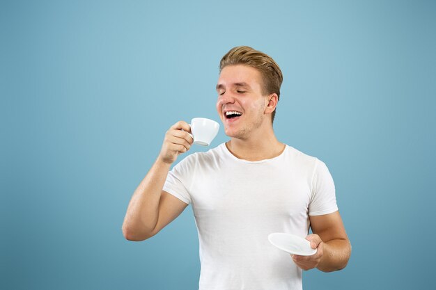 Caucasian young man's half-length portrait on blue studio background. Beautiful male model in shirt. Concept of human emotions, facial expression, sales, ad. Enjoying drinking coffee or tea.