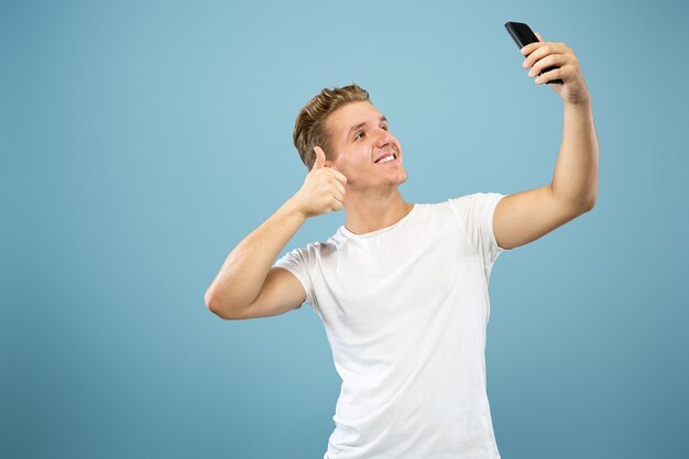 Caucasian young man's half-length portrait on blue studio background. Beautiful male model in shirt. Concept of human emotions, facial expression, sales, ad. Making selfie or content for vlog.