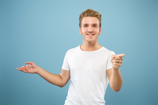 Caucasian young man's half-length portrait on blue studio background. Beautiful male model in shirt. Concept of human emotions, facial expression, sales, ad. Pointing and showing something.