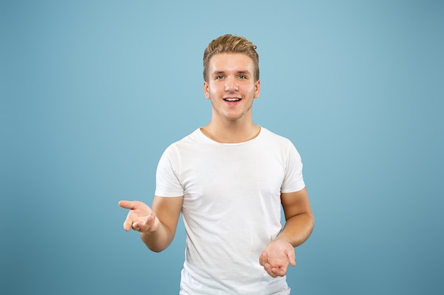 Caucasian young man's half-length portrait on blue studio background. Beautiful male model in shirt. Concept of human emotions, facial expression, sales, ad. Pointing and showing something.