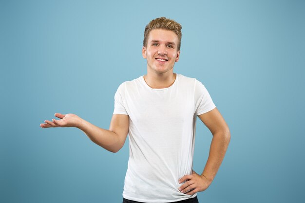 Caucasian young man's half-length portrait on blue studio background. Beautiful male model in shirt. Concept of human emotions, facial expression, sales, ad. Pointing and showing something.