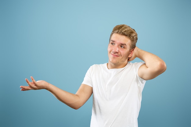 Caucasian young man's half-length portrait on blue studio background. Beautiful male model in shirt. Concept of human emotions, facial expression, sales, ad. Showing, looks uncertain, copyspace.