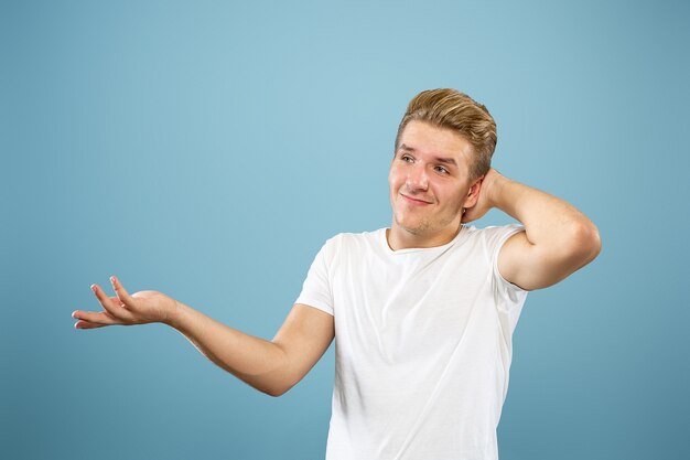 Caucasian young man's half-length portrait on blue studio background. Beautiful male model in shirt. Concept of human emotions, facial expression, sales, ad. Showing, looks uncertain, copyspace.