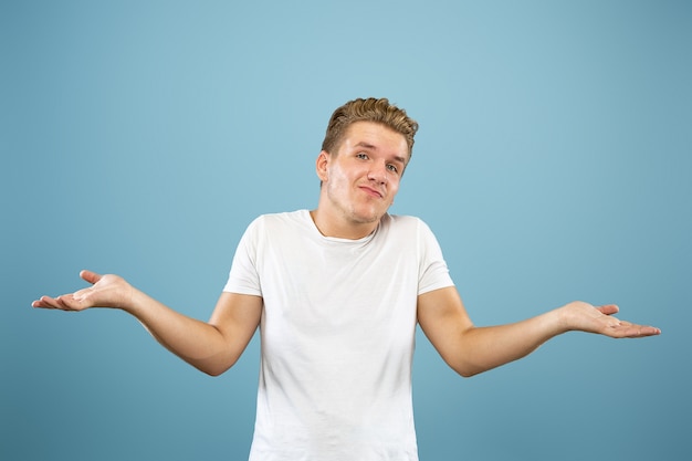 Caucasian young man's half-length portrait on blue studio background. Beautiful male model in shirt. Concept of human emotions, facial expression, sales, ad. Showing, looks uncertain, copyspace.