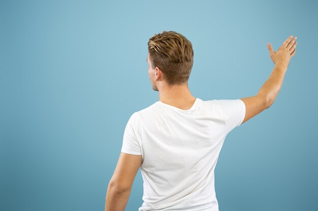 Caucasian young man's half-length portrait on blue studio background. Beautiful male model in shirt. Concept of human emotions, facial expression, sales, ad. Showing, pointing, copyspace.