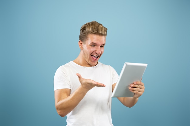 Caucasian young man's half-length portrait on blue studio background. Beautiful male model in shirt. Concept of human emotions, facial expression, sales, ad. Using tablet for vlog, selfie, study.