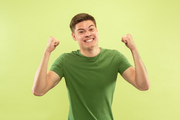 Caucasian young man's half-length portrait on green studio background. Beautiful male model in shirt. Concept of human emotions, facial expression, sales, ad. Crazy happy, celebrating, shocked.