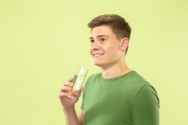 Caucasian young man's half-length portrait on green studio background. Beautiful male model in shirt. Concept of human emotions, facial expression, sales, ad. Enjoying drinking pure water.