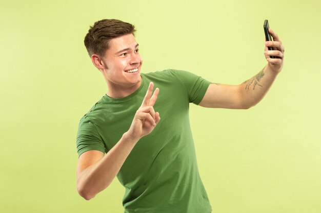 Caucasian young man's half-length portrait on green studio background. Beautiful male model in shirt. Concept of human emotions, facial expression, sales, ad. Making selfie or content for his vlog.