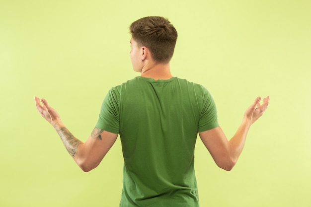 Caucasian young man's half-length portrait on green studio background. Beautiful male model in shirt. Concept of human emotions, facial expression, sales, ad. Showing and pointing something.