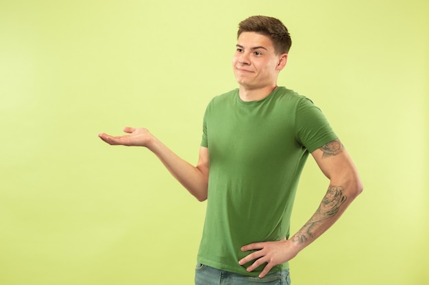 Caucasian young man's half-length portrait on green studio background. Beautiful male model in shirt. Concept of human emotions, facial expression, sales, ad. Uncertainty, doesn't know.