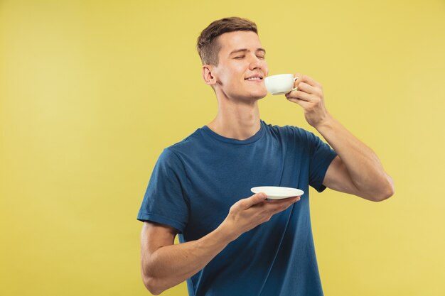 Caucasian young man's half-length portrait on yellow studio background. Beautiful male model in blue shirt. Concept of human emotions, facial expression. Enjoying drinking coffee or tea.
