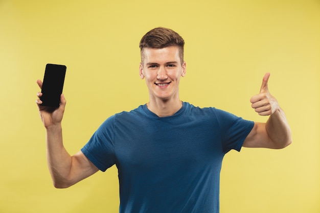 Caucasian young man's half-length portrait on yellow studio background. Beautiful male model in blue shirt. Concept of human emotions, facial expression. Holding phone and showing thumb up.
