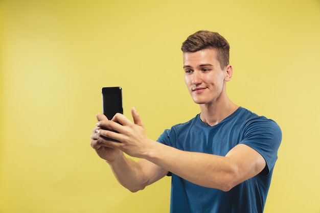 Caucasian young man's half-length portrait on yellow studio background. Beautiful male model in blue shirt. Concept of human emotions, facial expression. Making selfie or vlog, looks happy.