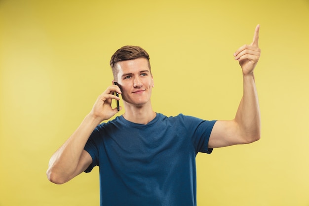 Caucasian young man's half-length portrait on yellow studio background. Beautiful male model in blue shirt. Concept of human emotions, facial expression. Talking on phone, have an idea.