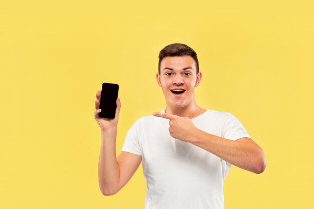 Поясной портрет кавказского молодого человека на желтом фоне студии. Красивая мужская модель в рубашке. Концепция человеческих эмоций, выражения лица, продаж, рекламы. Показываю экран телефона и улыбаюсь.