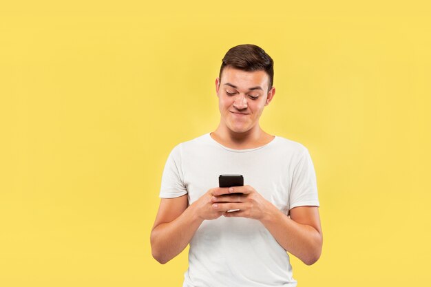 Caucasian young man's half-length portrait on yellow studio background. Beautiful male model in shirt. Concept of human emotions, facial expression, sales, ad. Using phone, looks crazy happy.