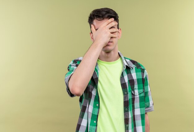 緑のシャツを着ている白人の若い男は、孤立した緑の壁に手で顔を覆った