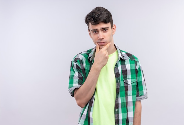 caucasian young man wearing green shirt put his hand on chin on isolated white wall