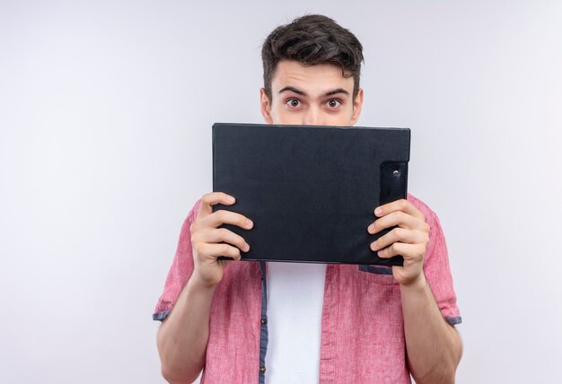 Caucasian young man wearing pink shirt covered face with clipboard on isolated white wall