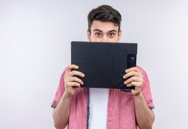 caucasian young man wearing pink shirt covered face with clipboard on isolated white wall