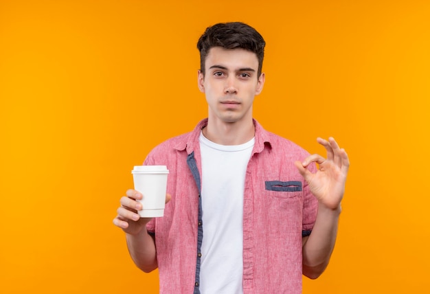 Кавказский молодой человек в розовой рубашке держит чашку кофе и показывает жест на изолированной оранжевой стене