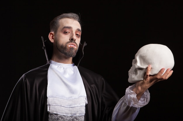Caucasian young man with scary face and dracula costume looking into the camera and holding a skull. Scary and dangerous man with vampire eyes.