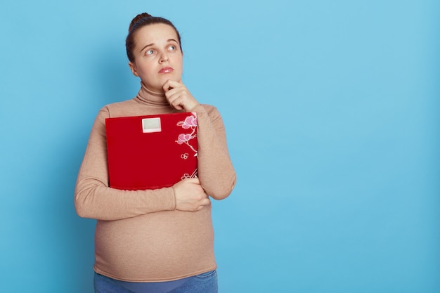 Free photo caucasian young woman in casual clothing posing isolated over blue wall looking aside with pensive expression, keeping hand on chin, future mother thinks about her weight, copy space