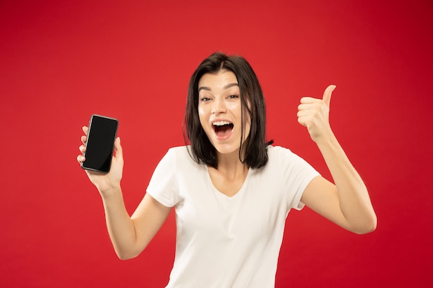 Caucasian young woman's half-length portrait on red studio background. Beautiful female model in white shirt. Concept of human emotions, facial expression, sales. Pointing with phone, looks happy.