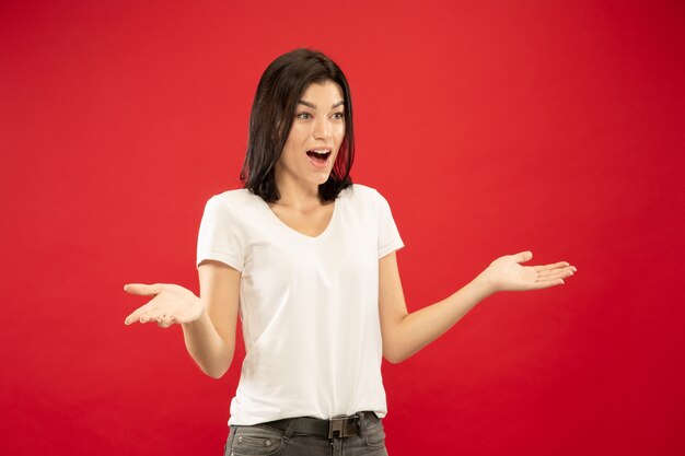 Caucasian young woman's half-length portrait on red studio background. Beautiful female model in white shirt. Concept of human emotions, facial expression. Showing something, looks astonished.