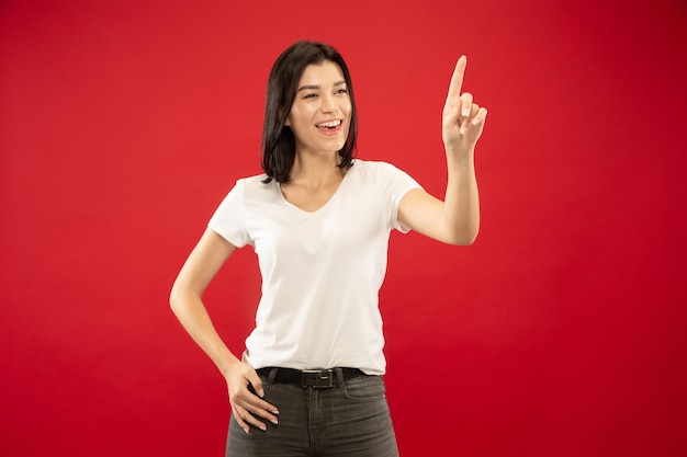 Caucasian young woman's half-length portrait on red studio background. Beautiful female model in white shirt. Concept of human emotions, facial expression. Touching empty search bar, copyspace.