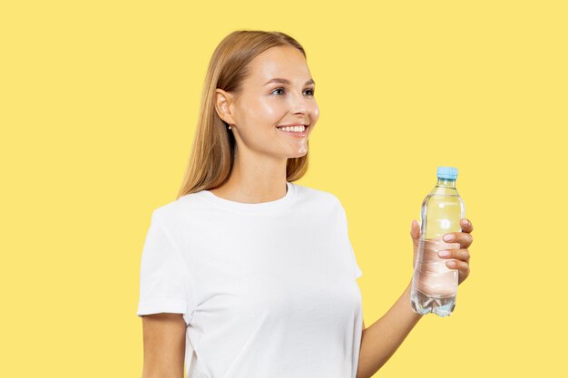 Caucasian young woman's half-length portrait on yellow studio background. Beautiful female model in white shirt. Concept of human emotions, facial expression. Drinking water from the bottle.